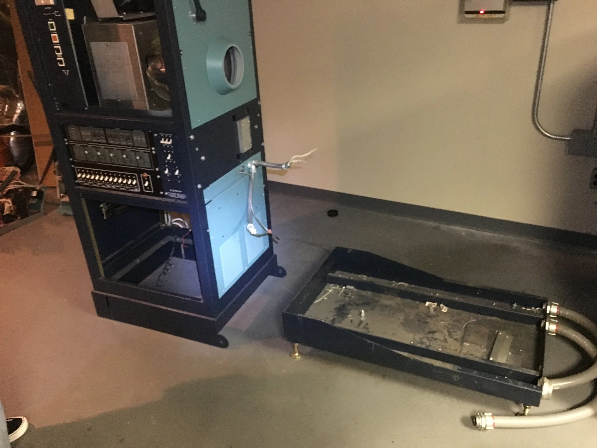 Disassembled electronic equipment with a partially opened black metal chassis and various electronic components inside, placed on a gray floor against a beige wall.