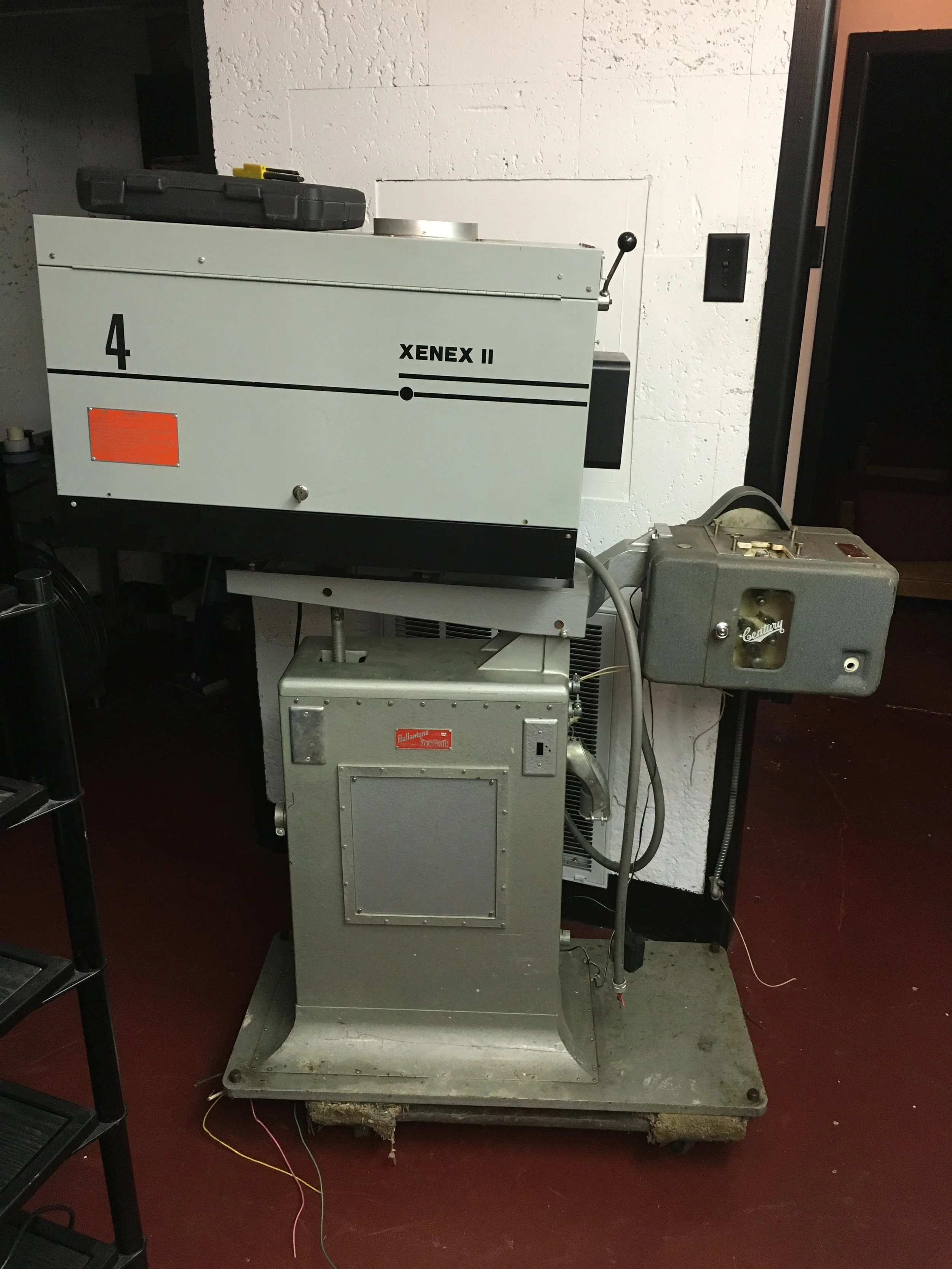 A laboratory machine labeled 'XENEX II' on the top panel, with a control arm, and placed on a rolling metal base. The background shows a white textured wall and a black switch.