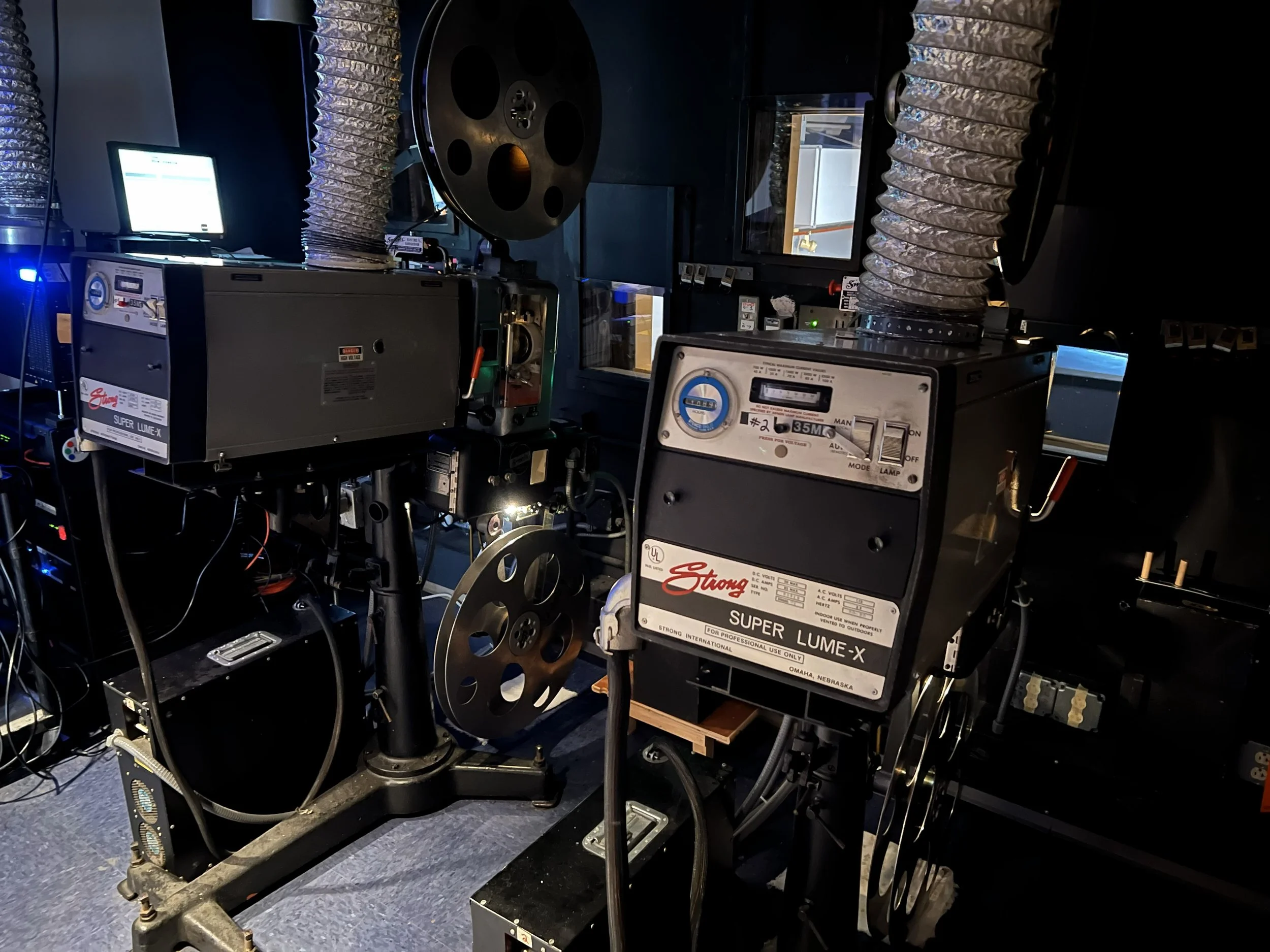 Close-up of film projectors with reels and ventilation hoses, set up in a dark room.