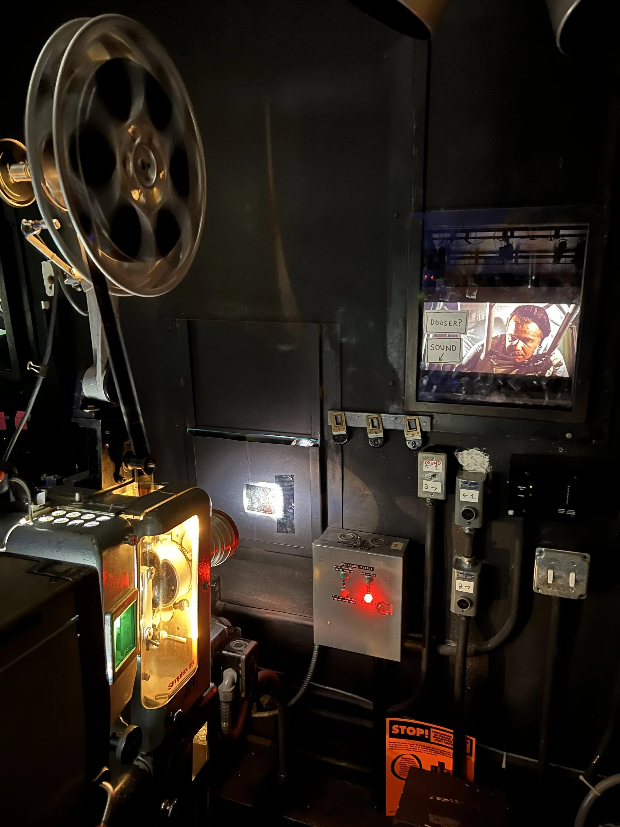 A vintage film projector in a dark room, with reels, a screen displaying a scene from a movie, and various control panels and switches.
