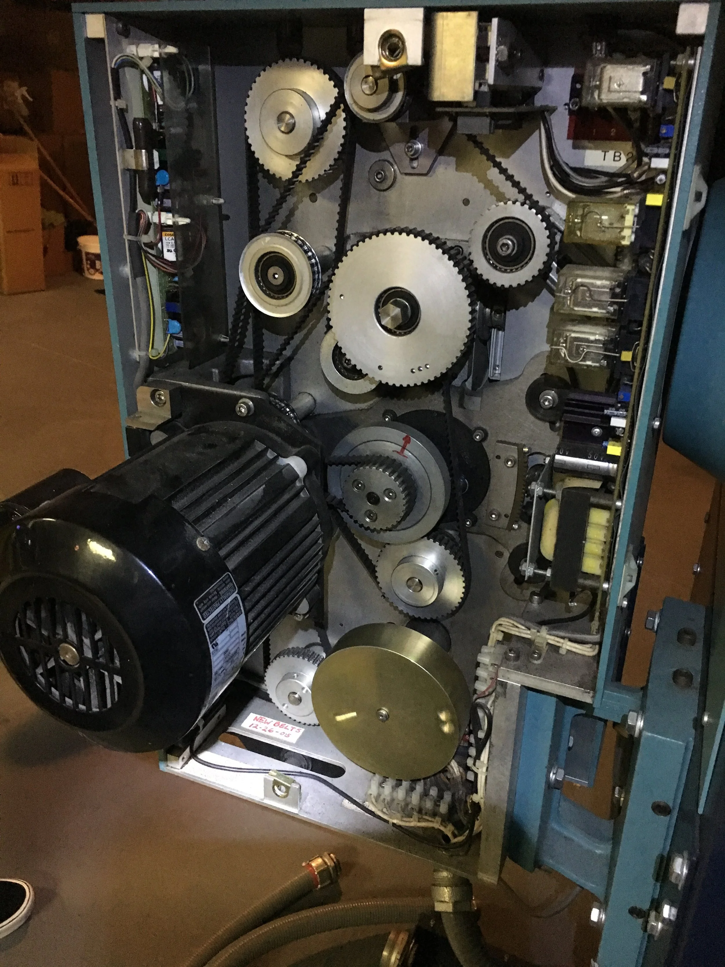 Open mechanical device with gears, belts, and motor components inside a metal frame, likely part of an industrial or mechanical machine.