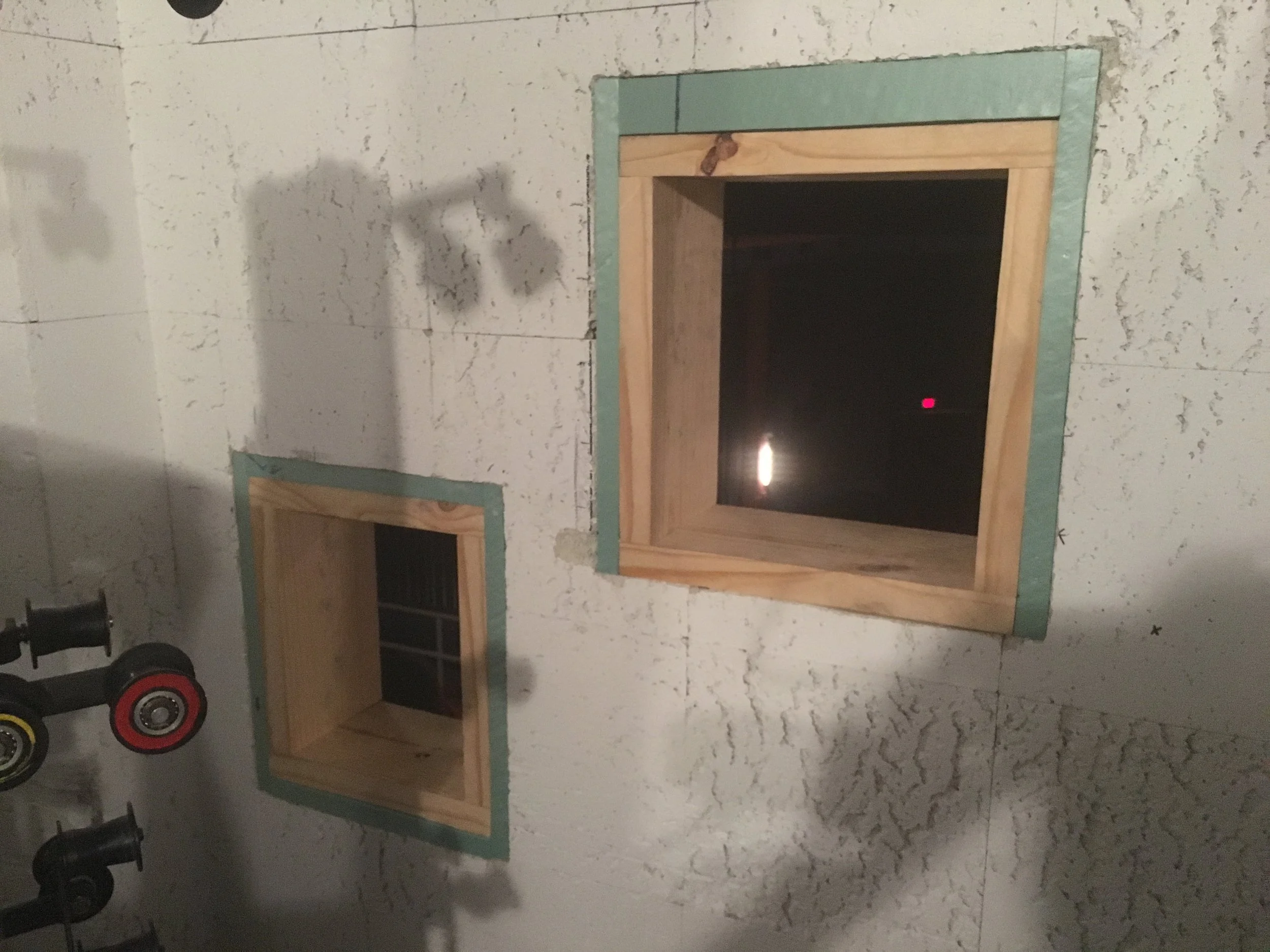 Two wooden window frames installed in a white wall with textured finish, one larger with a beveled border and the other smaller with a grille, during construction.