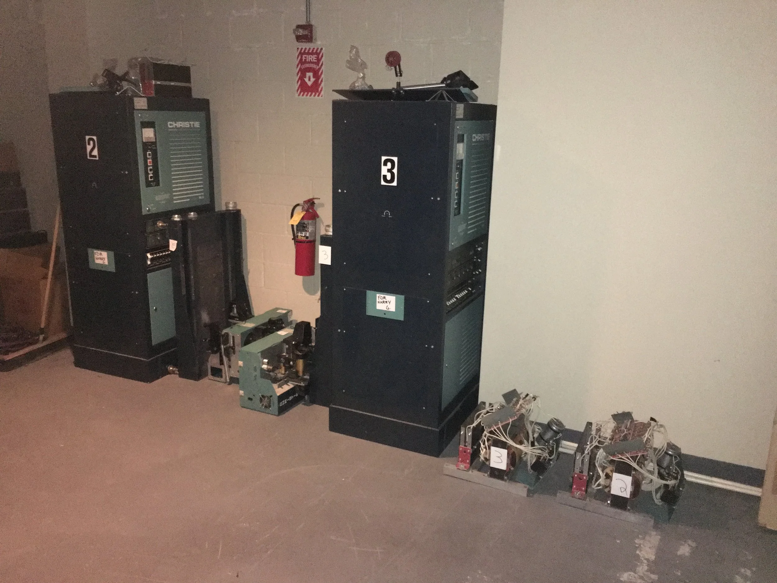Three large electrical or machinery cabinets are on the floor next to a beige wall. The cabinets are dark-colored with control panels and labels. To the right on the floor, smaller mechanical components with wires are arranged. A fire extinguisher ha