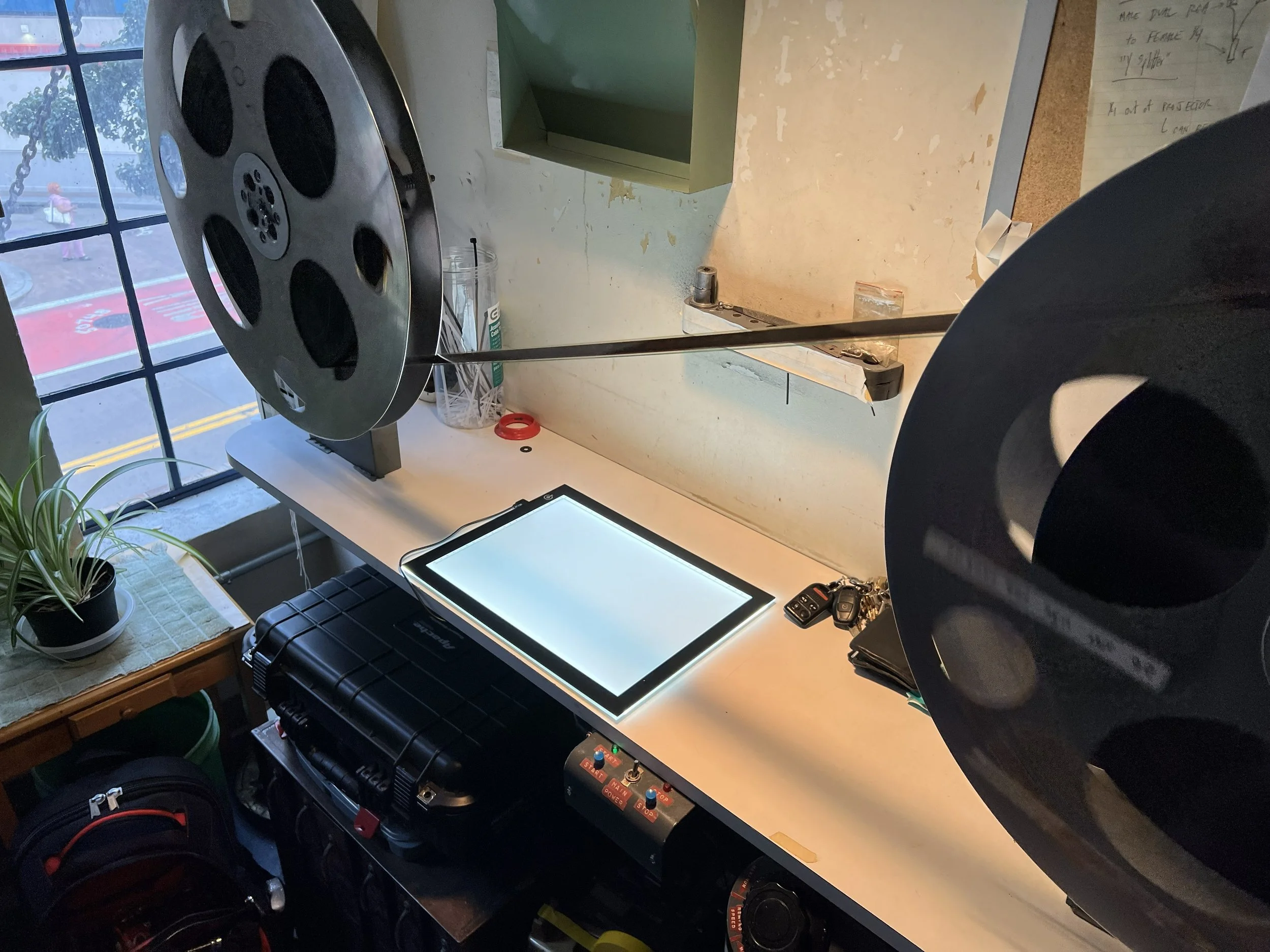 A workspace with two film projectors hanging from the ceiling, a digital tablet, car keys, and a control panel on the table. There's a potted plant on a small side table near a window, and the window shows a street outside.
