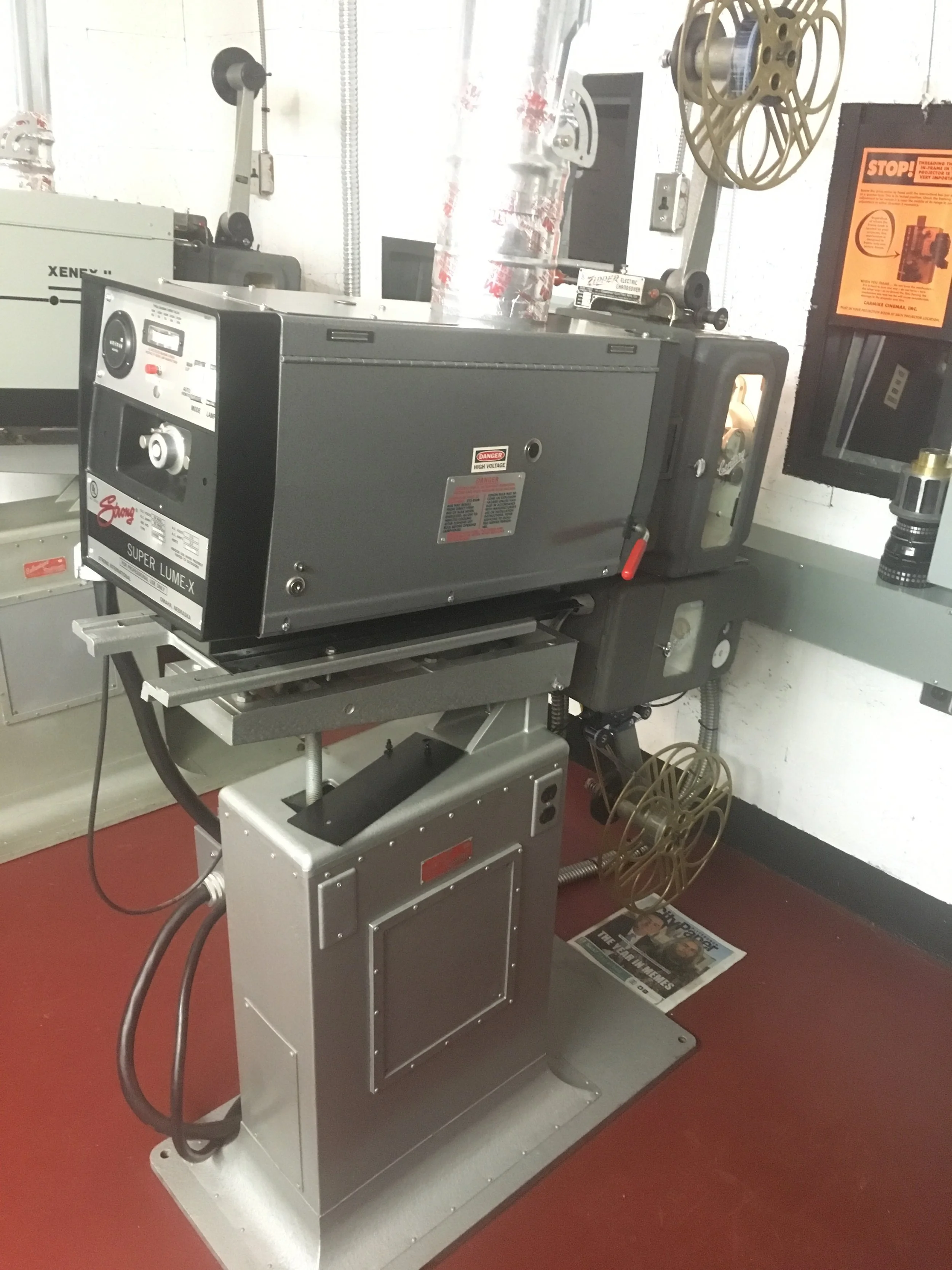 A laser engraving machine on a metal stand in a workshop. The machine has a gray body with control knobs and a digital display, and a large round component with a belt wheel. There's a safety window on the side, and a newspaper is on the floor beneat