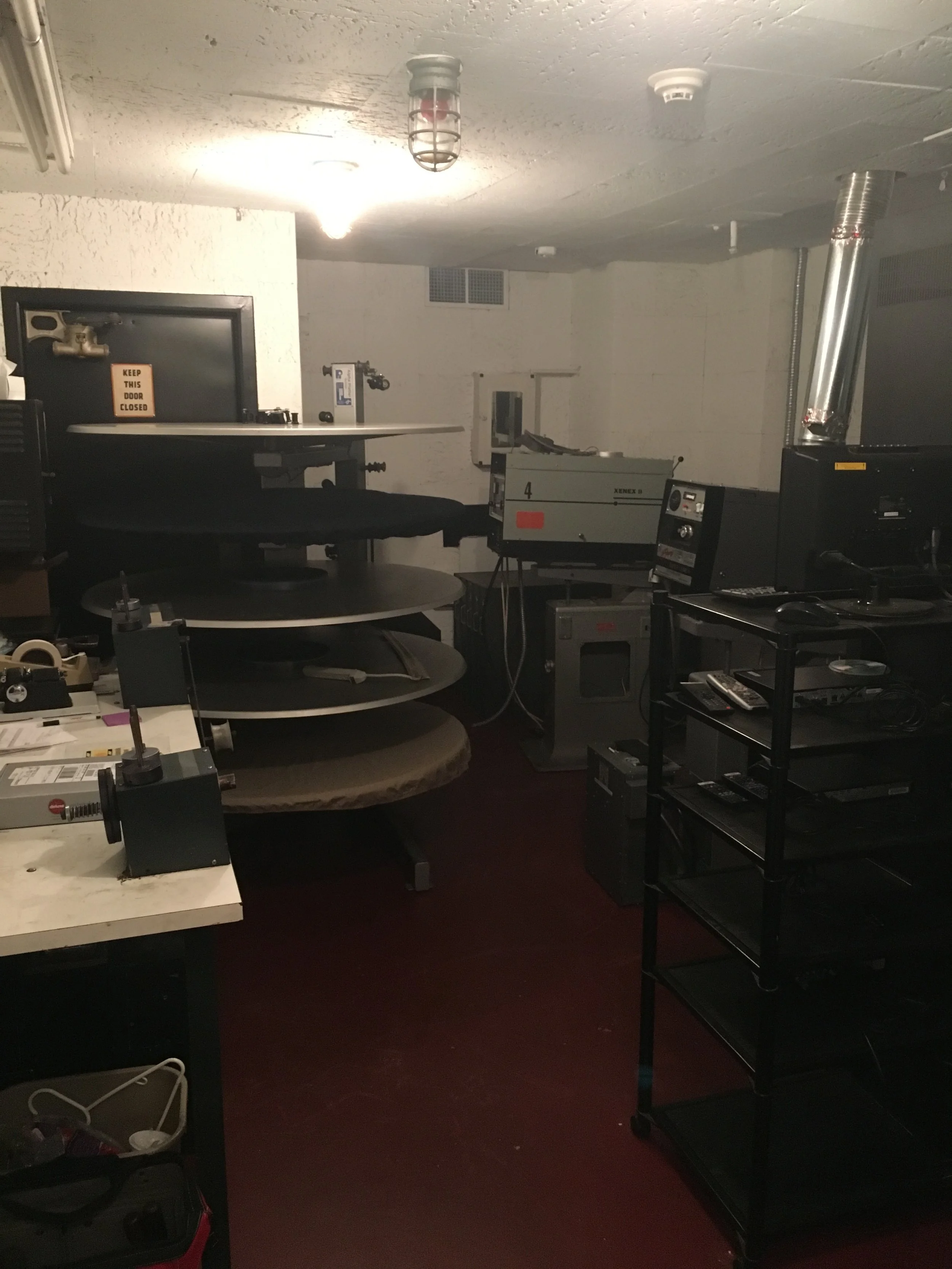 Room with printing or manufacturing equipment, including large circular machines, shelves with tools and supplies, and control panels.