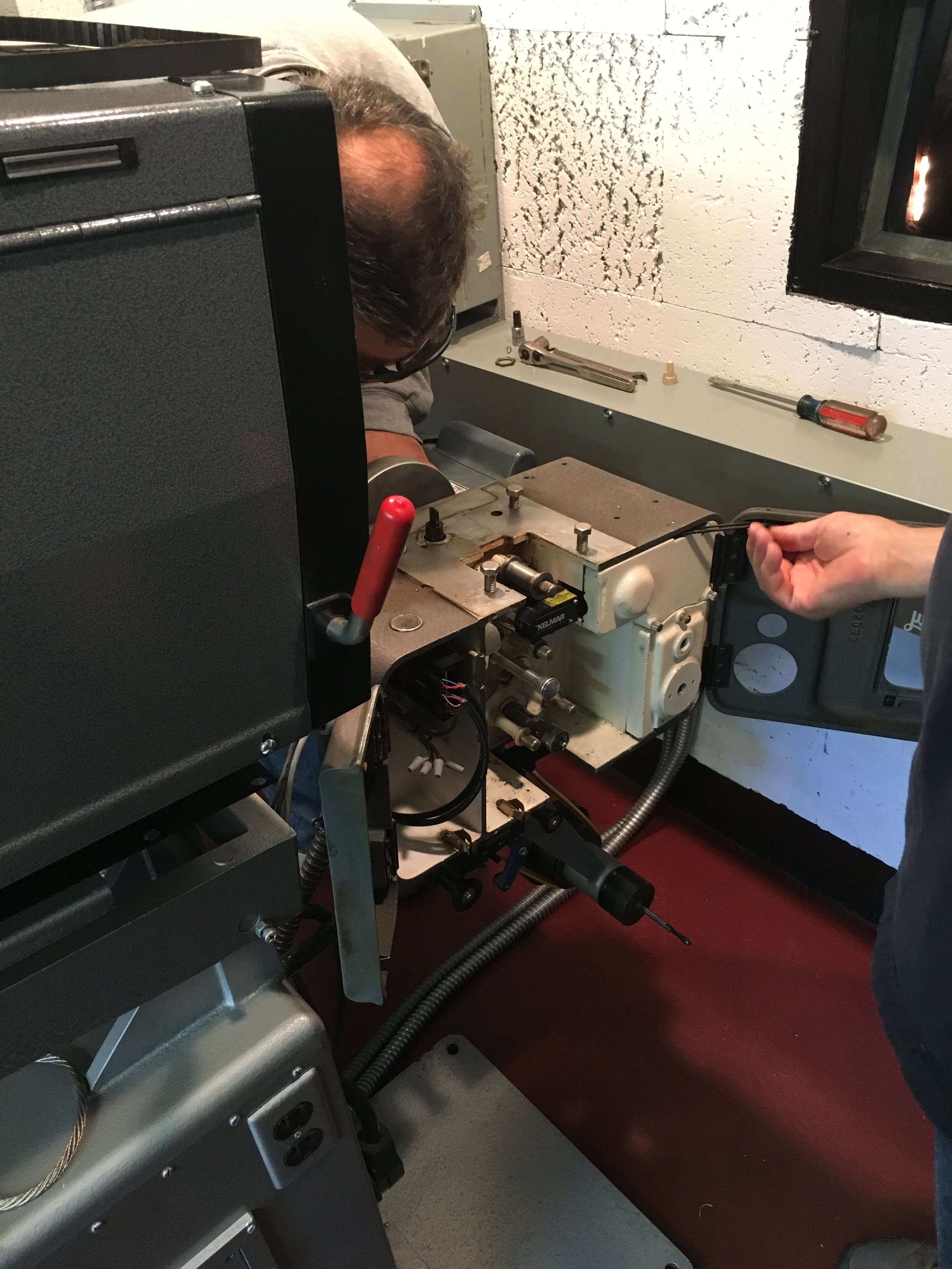 Two people working on a machine, one person is partially visible on the right side holding a tool, and another person is mostly obscured, peering over the machine with only part of their head visible. The setting appears to be a workshop or industria