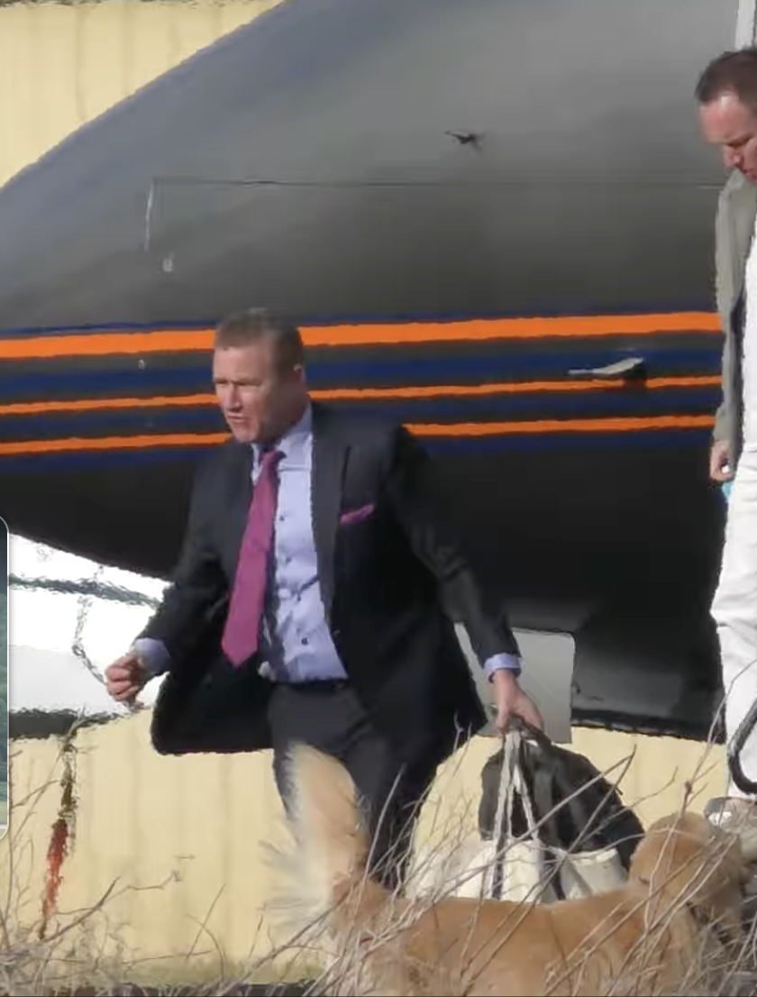 Man in a suit carrying a bag and a dog with two other men walking near a black boat shaped object with orange and blue stripes.