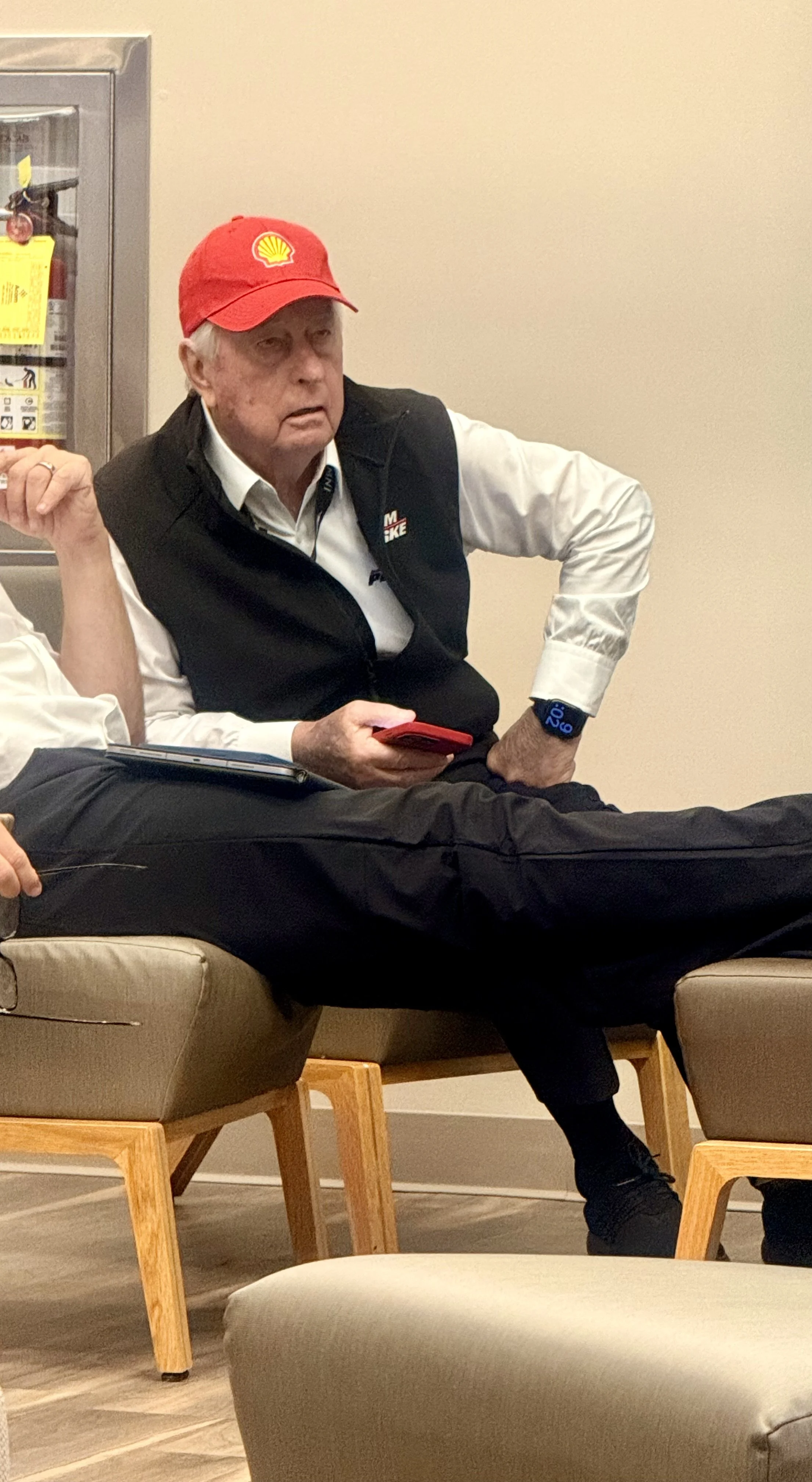 A man sitting on a beige chair, wearing a red Shell cap and a black vest over a white shirt, holding a red phone, with a smartwatch on his wrist.