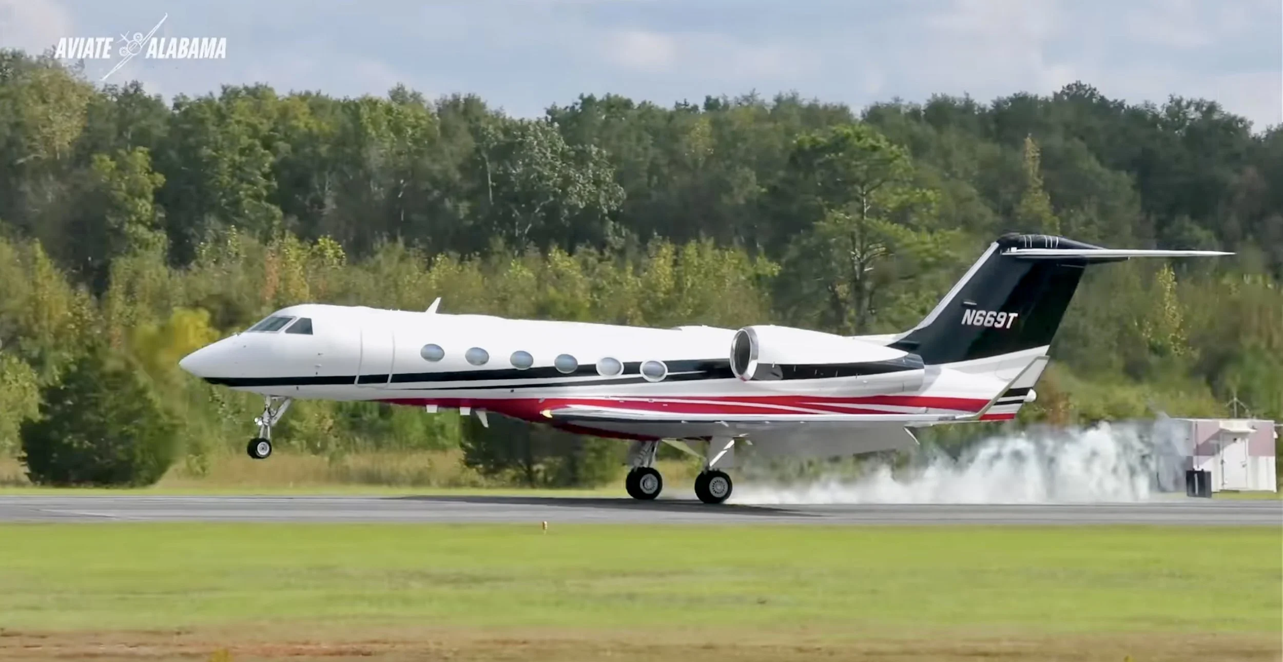 A white private jet with black and red accents taking off from a runway with clouds and trees in the background.