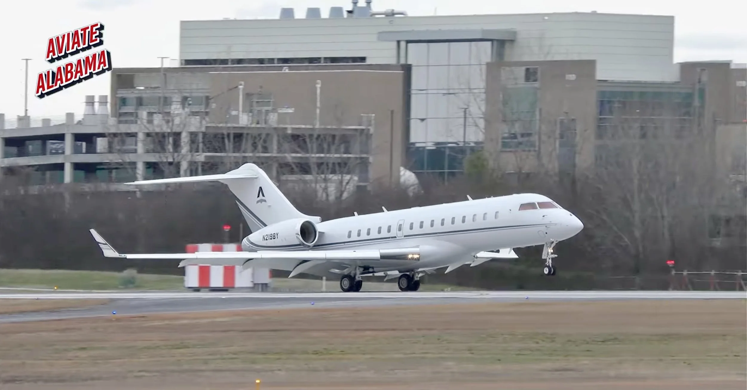 A white private jet taking off from an airport runway with a cityscape in the background and a sign that reads 'Aviate Alabama'.