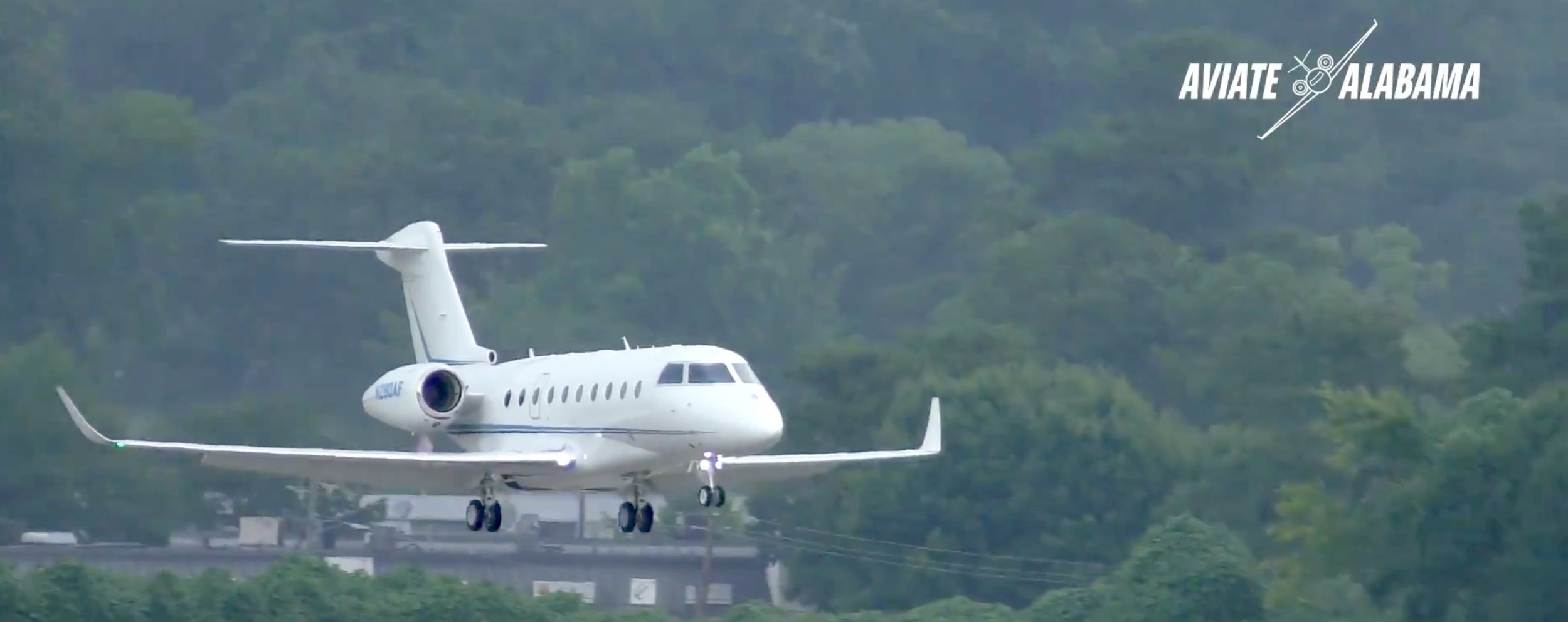A white private jet aircraft flying low over green trees and a highway with mountains in the background, with 'Aviate Alabama' logo in the top right corner.