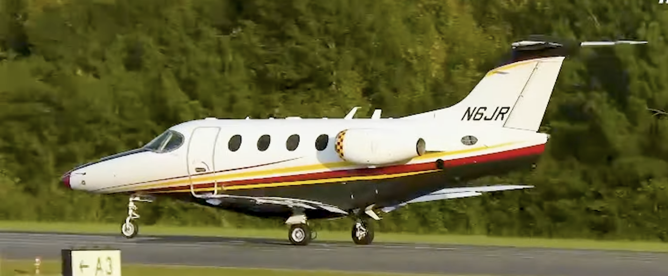 A small white private jet with yellow, red, and black stripes landing on a runway, surrounded by green trees.