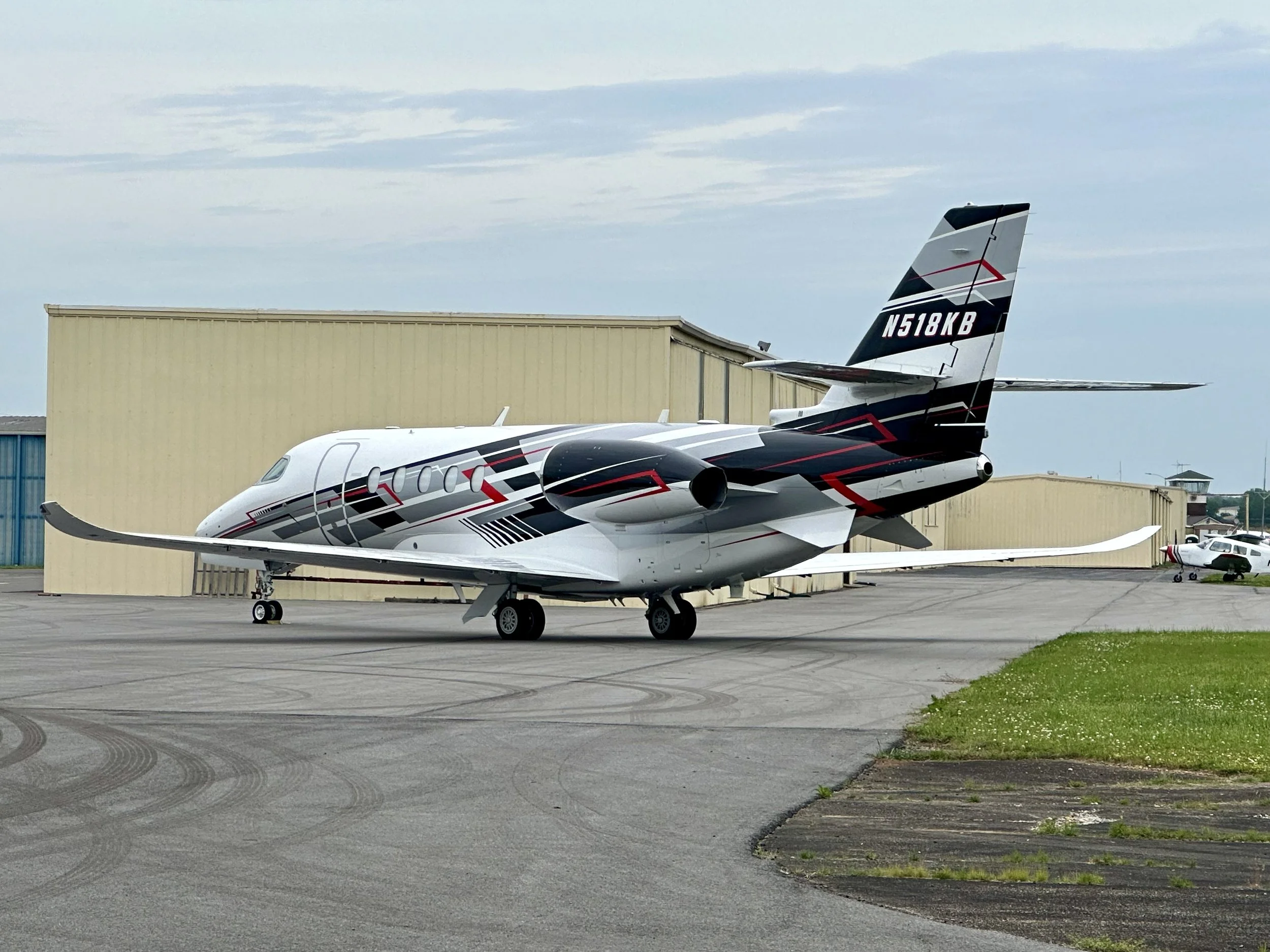 A small private jet with black, white, and red exterior design parked on an airport tarmac near a beige hangar, with a clear sky overhead.
