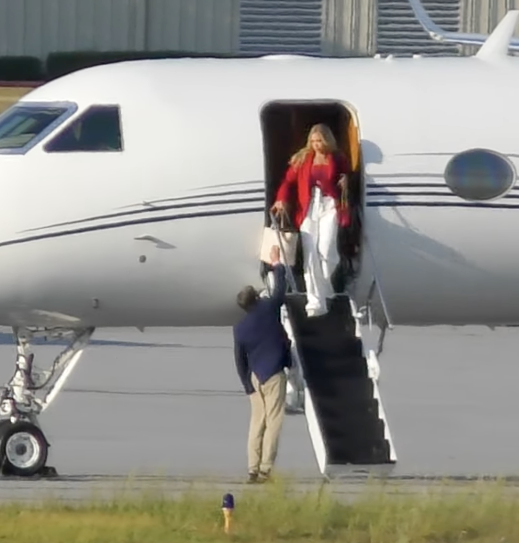 A woman in a red jacket and white pants exiting a private jet via the stairs, while a man in a blue jacket and beige pants stands on the ground near the steps.