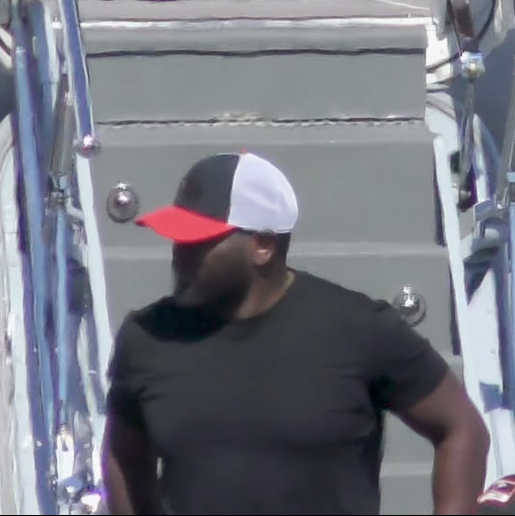 Person with long hair wearing a black T-shirt and a tricolored cap, standing near a decorated structure.