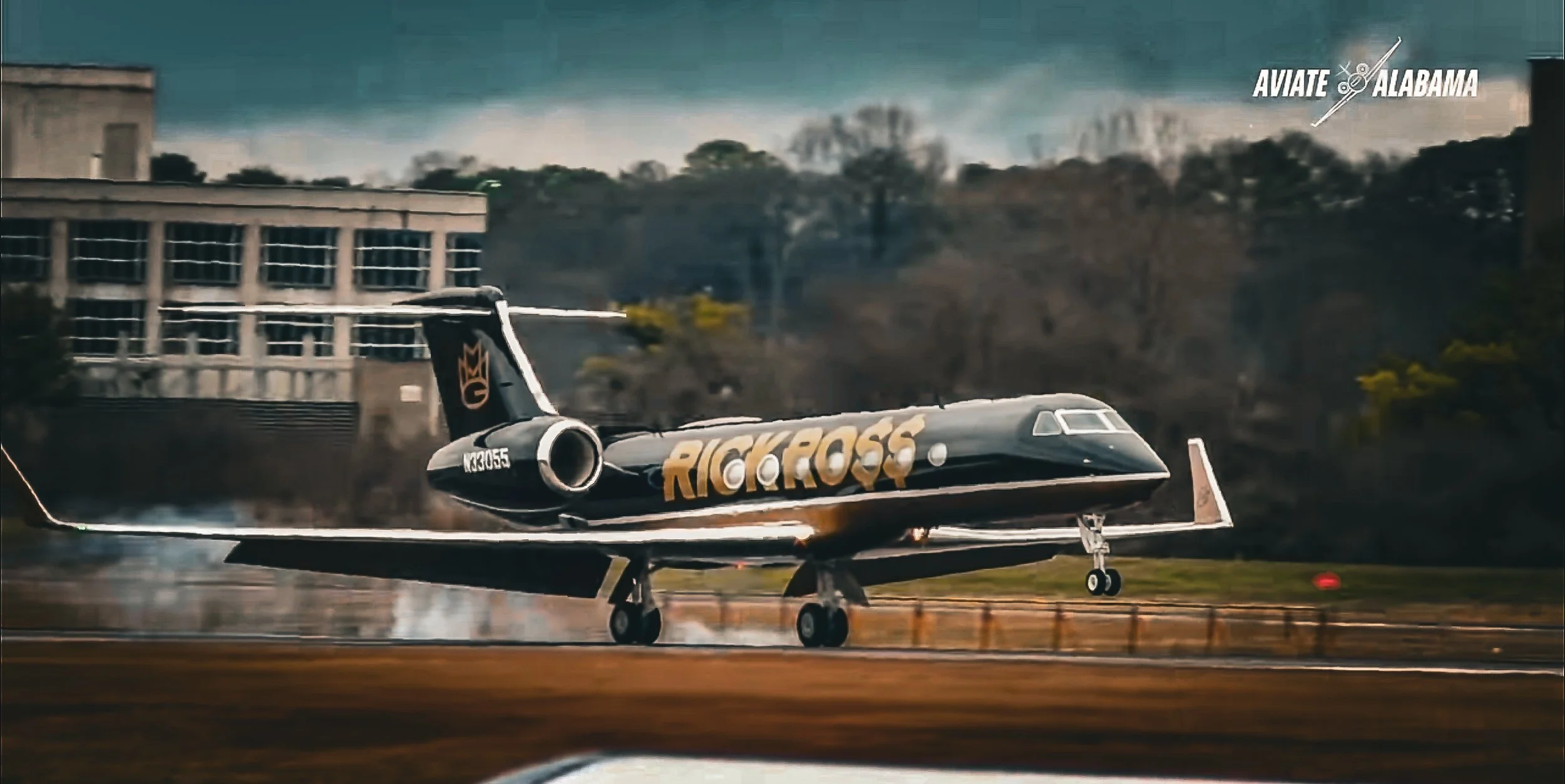 A stunt plane with branding 'Rick Ross' on the side is taking off from a runway, with smoke or vapor trailing behind it. The scene is set on a cloudy day in Alabama, as indicated by the 'Aviate Alabama' logo in the top right corner.