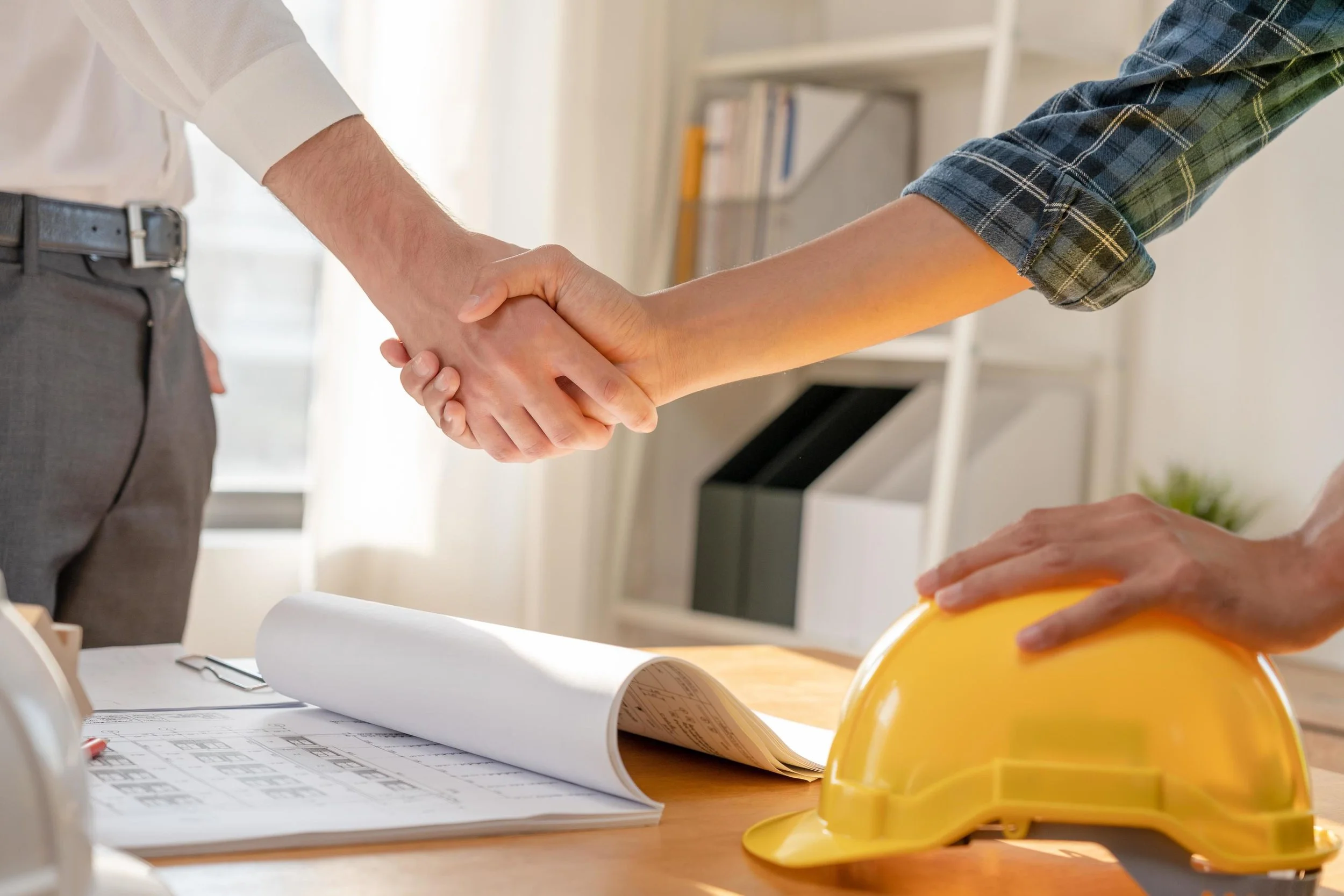 Two people shake hands over a table with architectural plans, a yellow safety helmet, and a clipboard in an office setting.