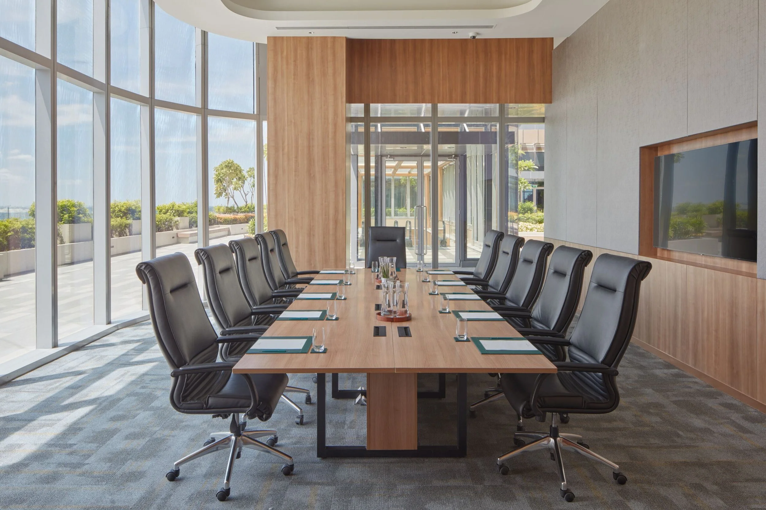 Empty conference room with a long wooden table, black leather chairs, large windows with outside view, and a wall-mounted TV.