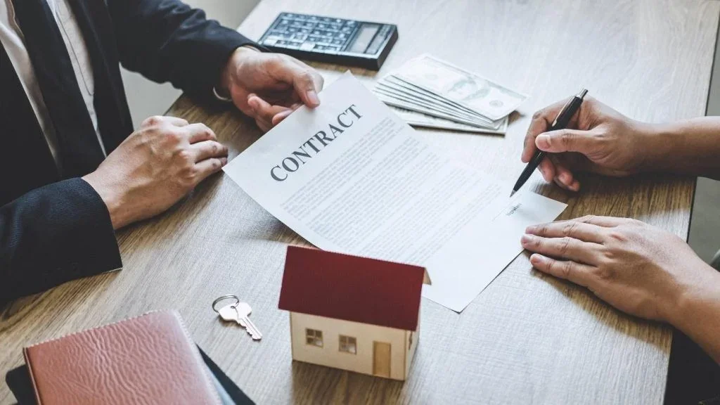Two people signing a contract at a desk with a small model house, keys, money, and a calculator.
