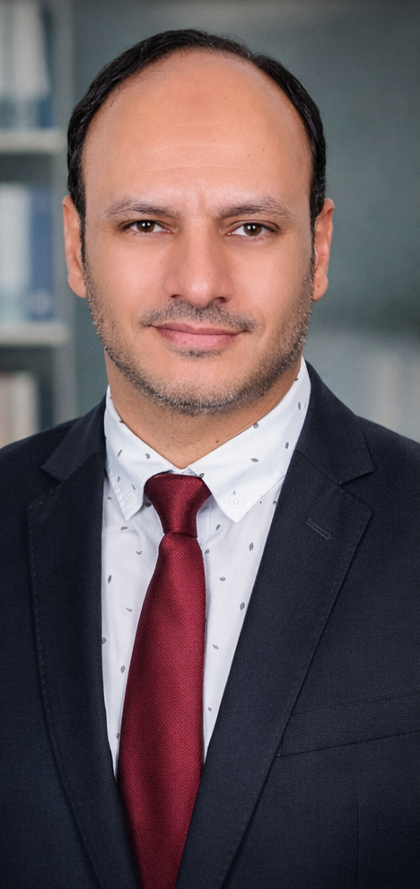 A professional man in a dark suit, white shirt with small patterns, and red tie, standing in an office setting with blurred bookshelves in the background.