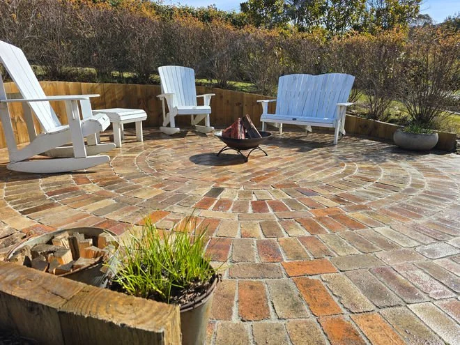Patio with white Adirondack chairs around a fire pit, brick pavers in a circular pattern, and potted plants, with trees and bushes in the background.