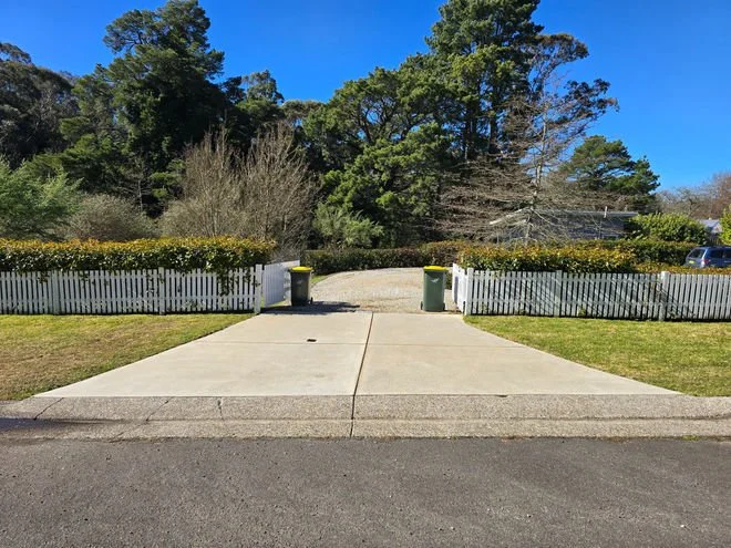 View of a driveway with two garbage bins, white picket fence on each side, leading to a park-like area with trees and blue sky.