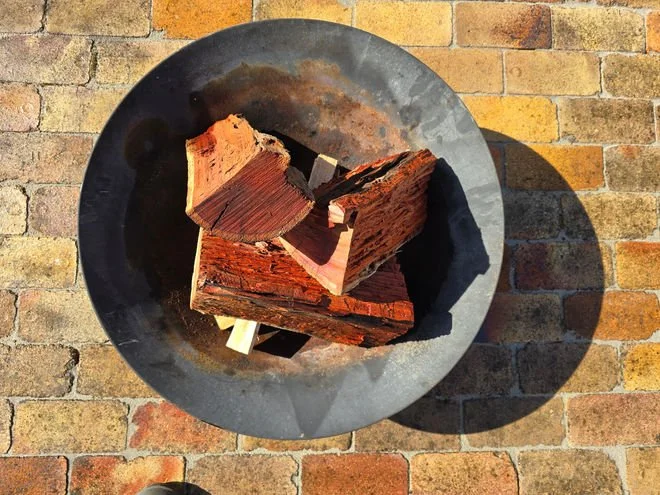 Charred wood pieces in a round black fire pit on a brick patio, with a shadow cast on the bricks.