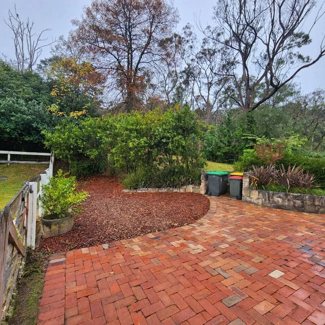 A brick patio in a backyard with a garden bed bordered by a brick wall and a variety of plants. Two trash bins are placed on the brick pathway. In the background, there are trees with some leaves changing color, indicating fall.