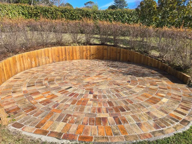 Circular brick patio with a wooden retaining wall and garden bushes in the background.