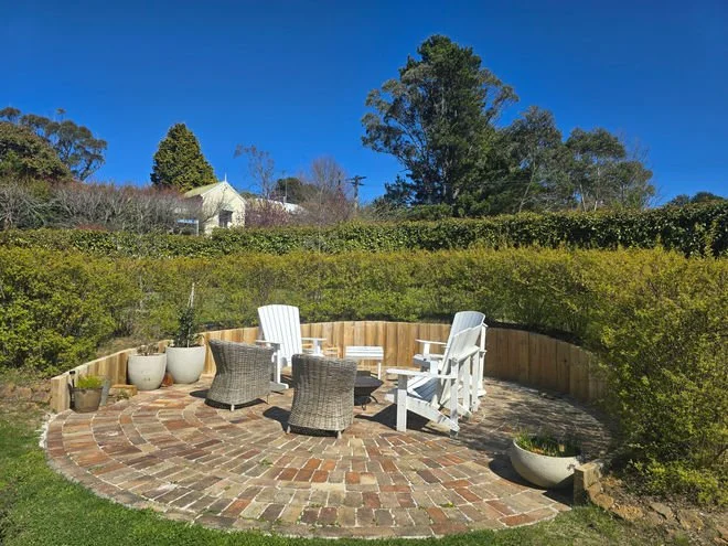 Outdoor patio with brick flooring, surrounded by a curved wooden fence and green bushes, featuring white and wicker chairs, potted plants, and a background of trees and houses under a clear blue sky.