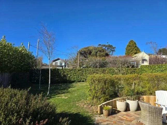 A backyard garden with a grassy slope, shrubs, trees, a patio area with outdoor chairs, and houses in the background under a clear blue sky.