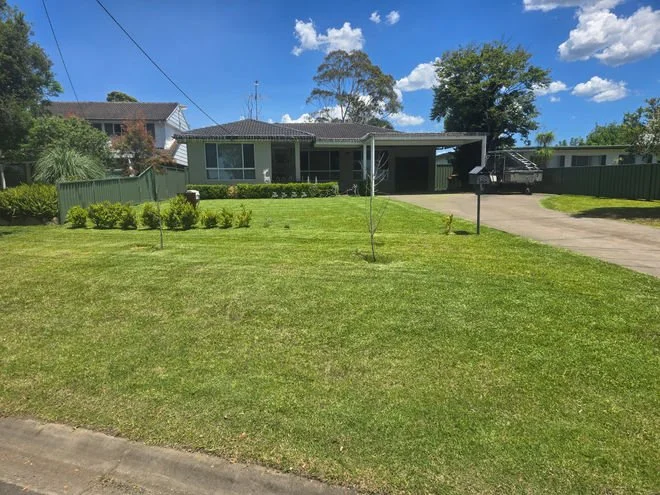 Front yard of a house with a well-maintained lawn, small trees, bushes, a concrete driveway, a carport, and a two-story house in the background under a blue sky with clouds.