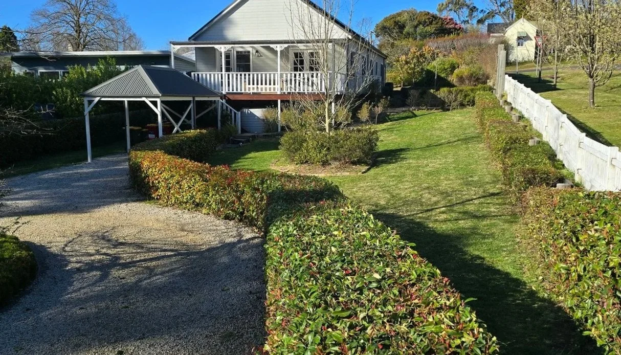 A white house with a porch and a balcony, surrounded by a well-manicured lawn, bushes, and trees. There is a gravel driveway leading up to a small covered structure near the house and a white fence on the right side of the lawn.