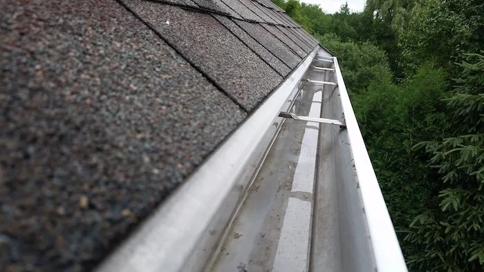 Close-up of a metal gutter on a house roof with shingles and green trees in the background.