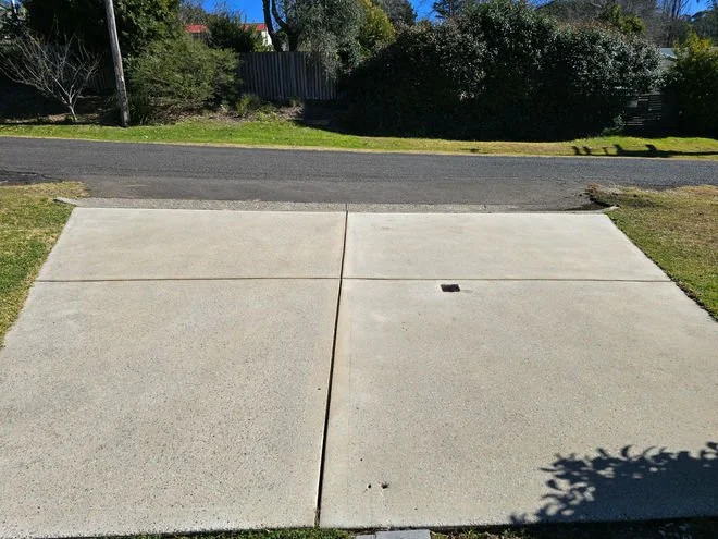A suburban driveway made of concrete slabs leading to a street with trees and bushes in the background.