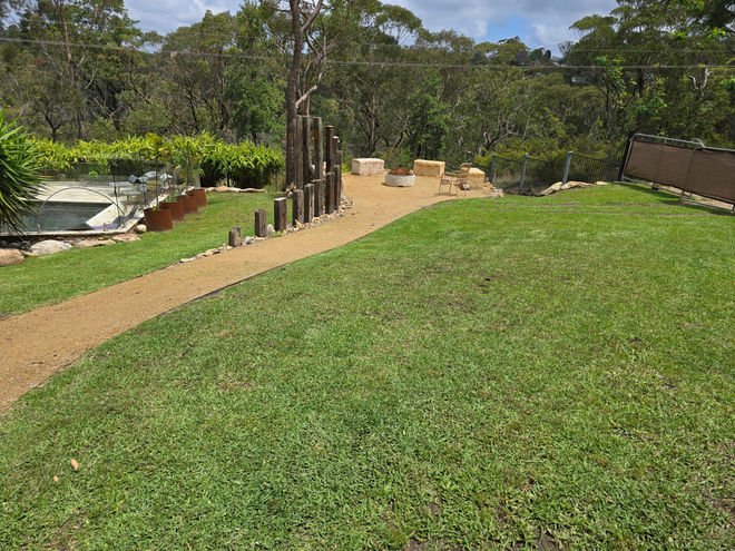 A backyard with a grassy lawn, a dirt pathway, and various stone and wood decorations.