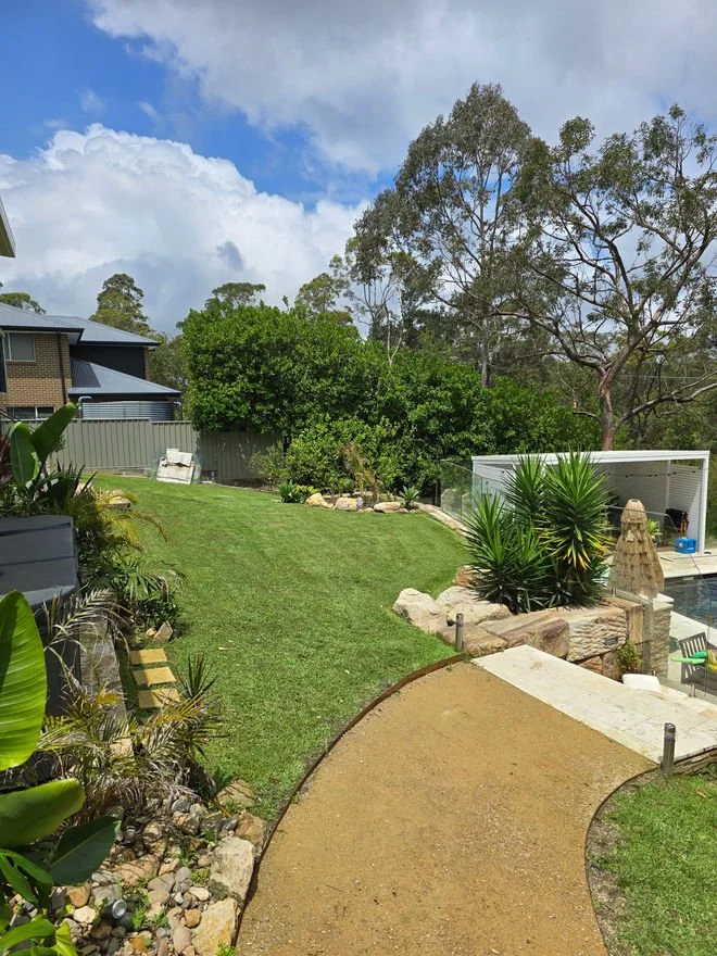 A backyard with a curved dirt pathway, green grass, various plants and trees, a cream-colored outdoor shelter, and a neighboring house in the background under a partly cloudy sky.