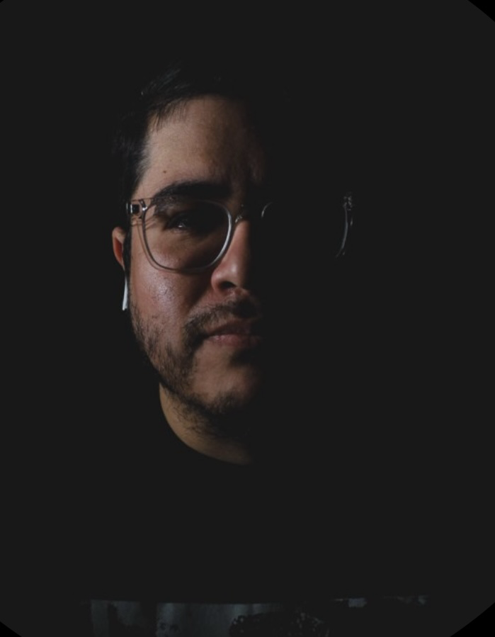 A man with glasses and facial hair shown in a dark setting with minimal lighting highlighting one side of his face.