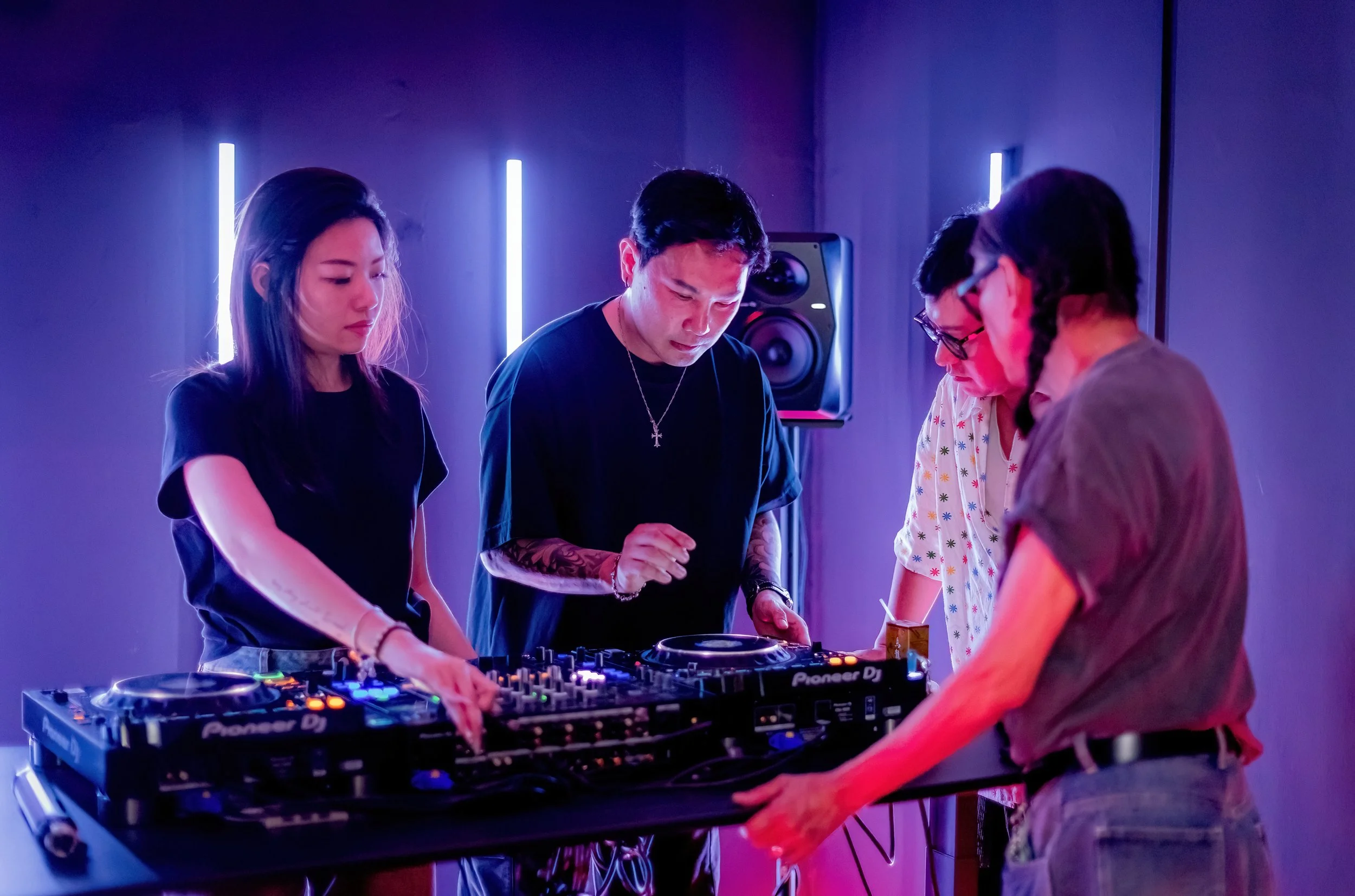 Four people gather around DJ equipment in a dimly lit room with vertical light bars, engaged in music mixing.