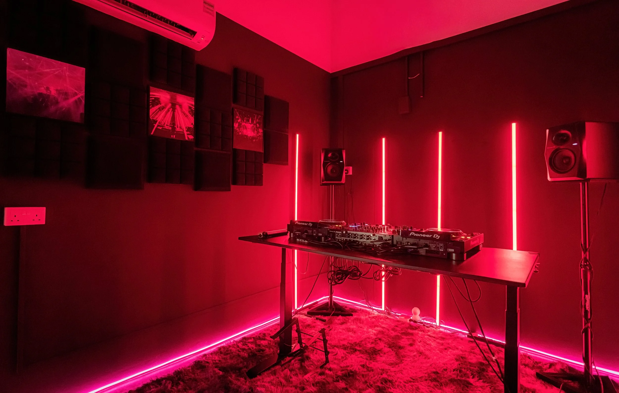 A DJ setup in a room illuminated by red LED lights, featuring a DJ mixer and turntables on a table, surrounded by speakers, with a patterned carpet on the floor and sound-absorbing panels on the walls.