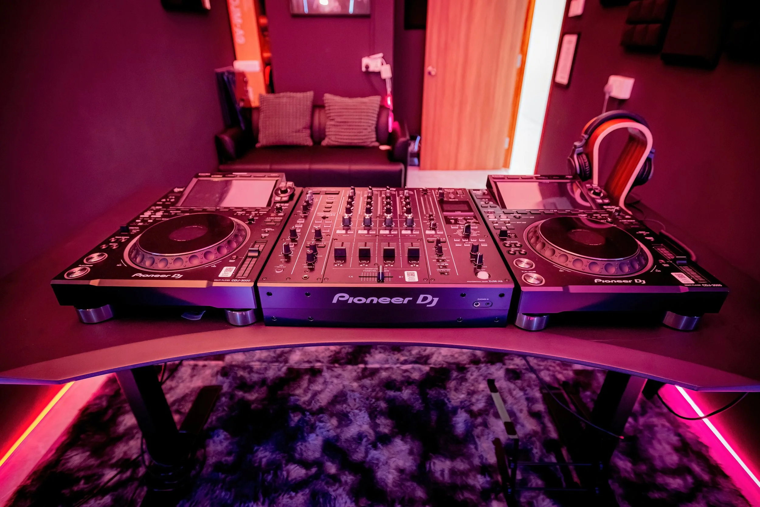 DJ equipment set up on a table with speakers, headphones, and control panel, in a room with purple walls and a couch with pillows in the background, illuminated with pink and purple lighting.