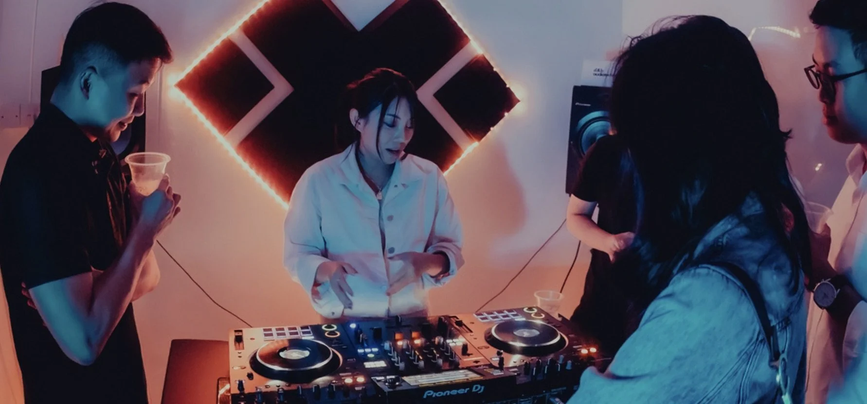 A female DJ in a white shirt DJing at a party with friends. A group of young people gathered around, some holding drinks, in a dimly lit room with red neon lights and sound equipment.