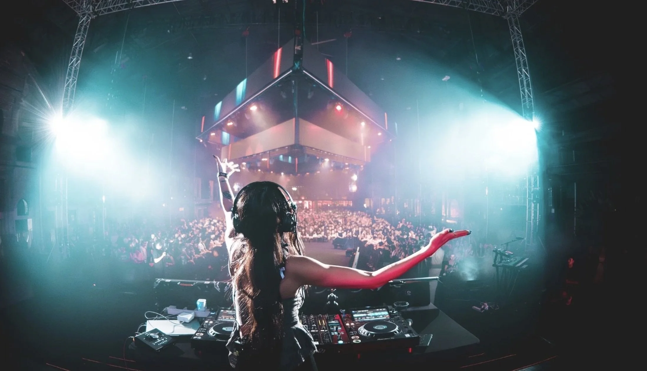 A DJ performing at a nightclub, with a large audience in front. The DJ is wearing headphones and has one arm raised. The scene is illuminated with colorful stage lights and spotlights creating a vibrant atmosphere.