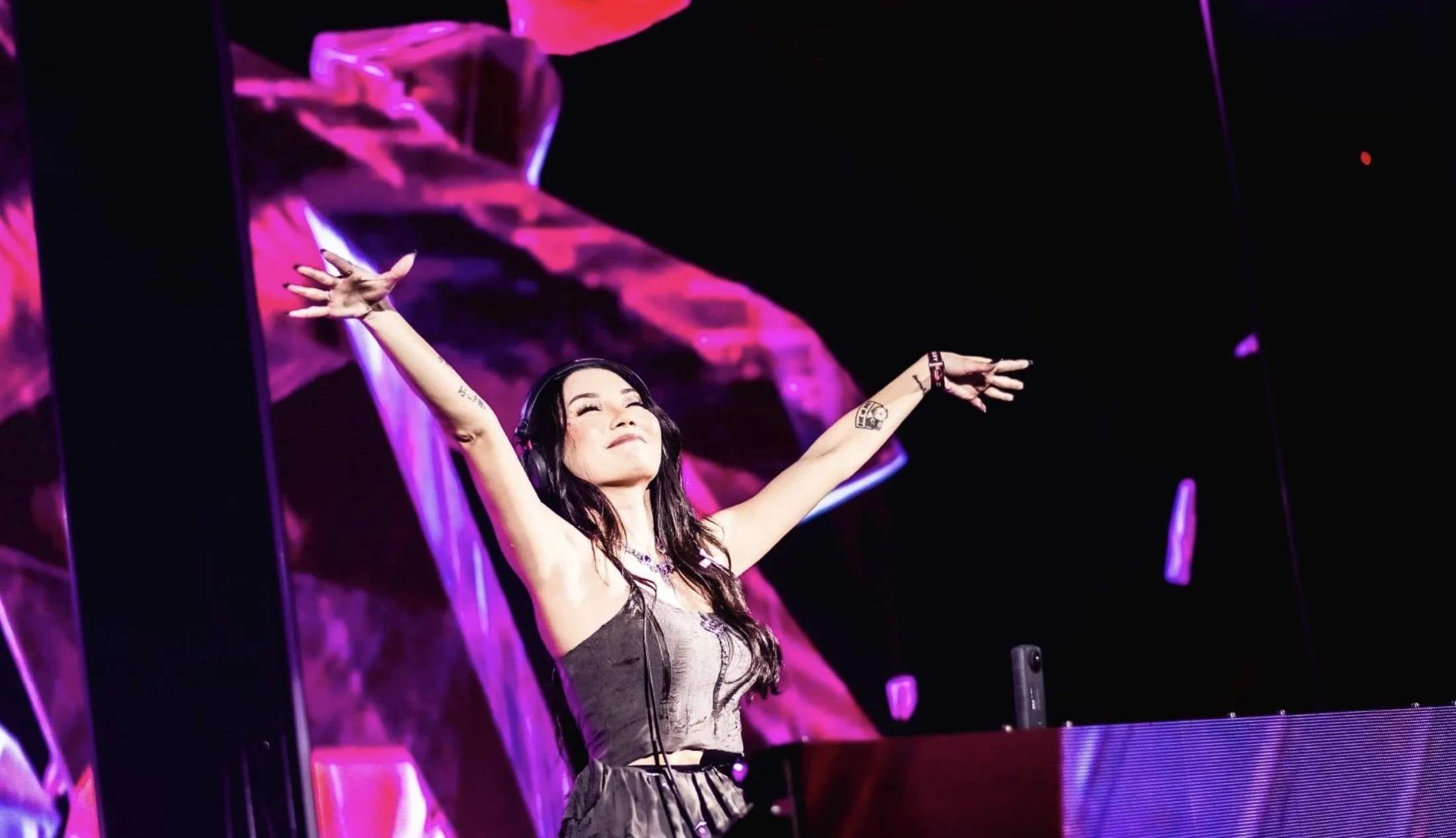 A woman with long dark hair, wearing headphones and a black dress, stands with arms outstretched in front of a DJ booth with purple and pink lighting effects.