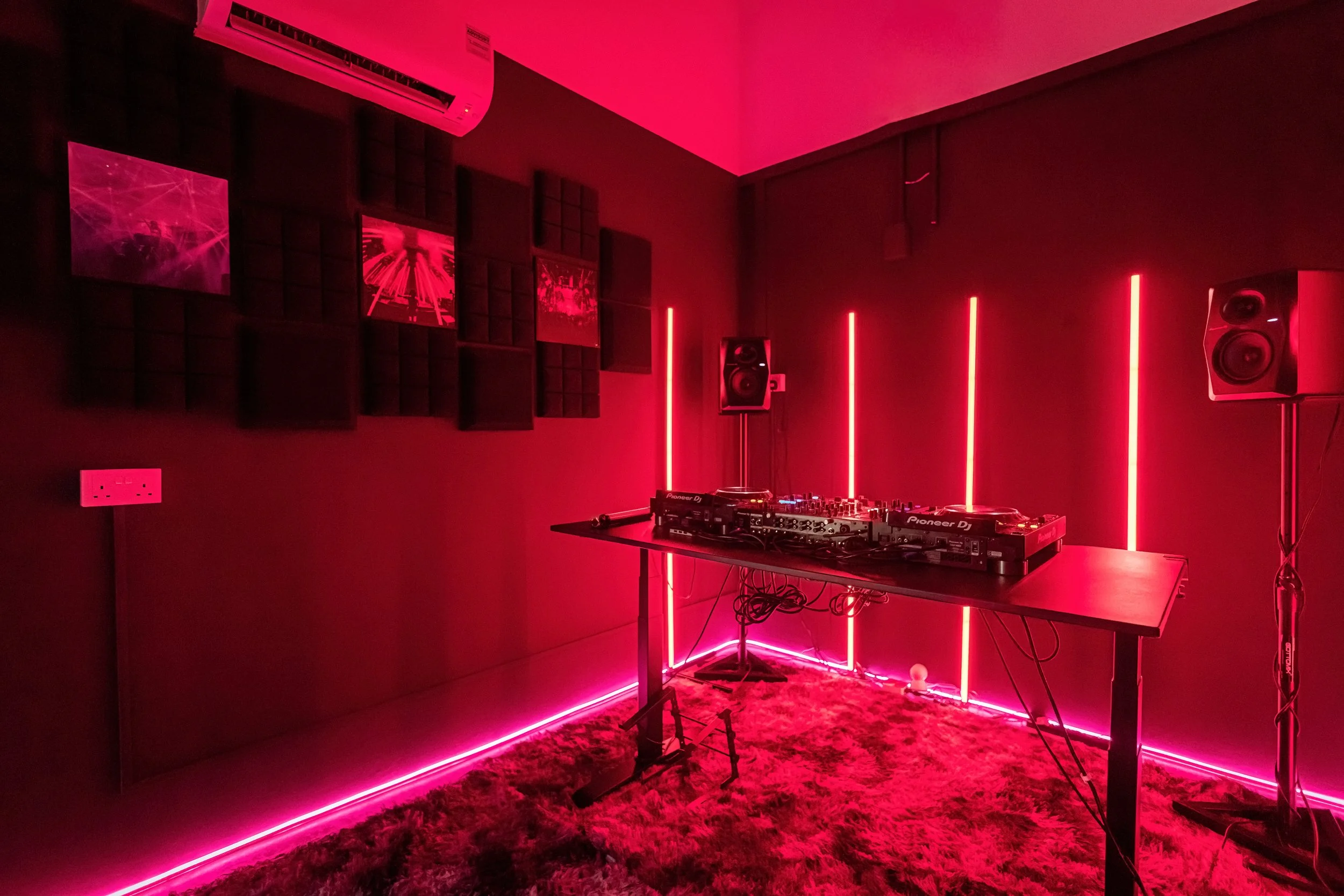 A DJ setup in a dimly lit room with pink neon lighting, speakers, DJ controller on a table, framed pictures on the wall, and a textured carpet.
