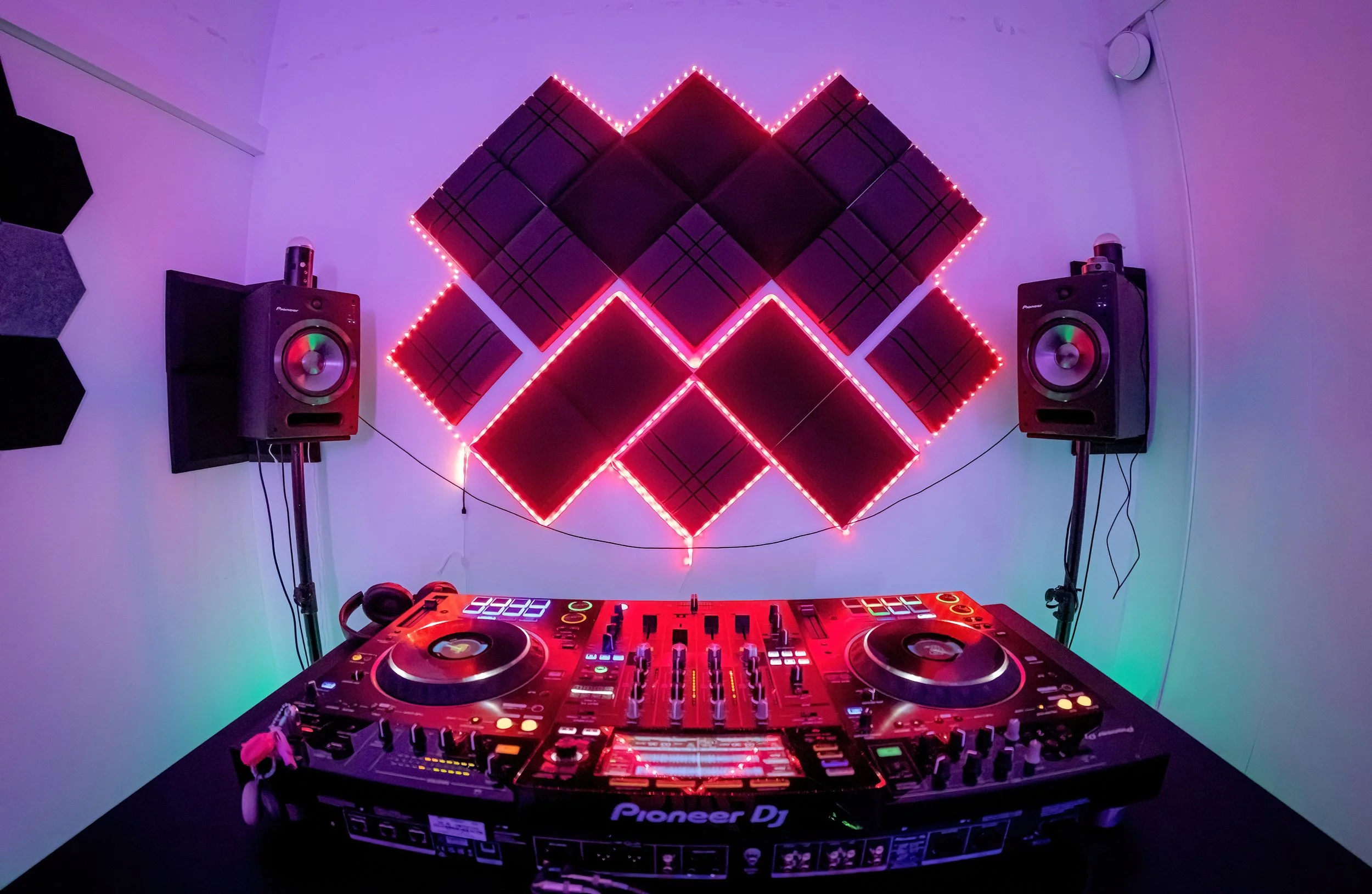 DJ setup with Pioneer DJ controller, dual speakers, and acoustic foam wall panels with pink and purple LED lighting in a music studio or club environment.
