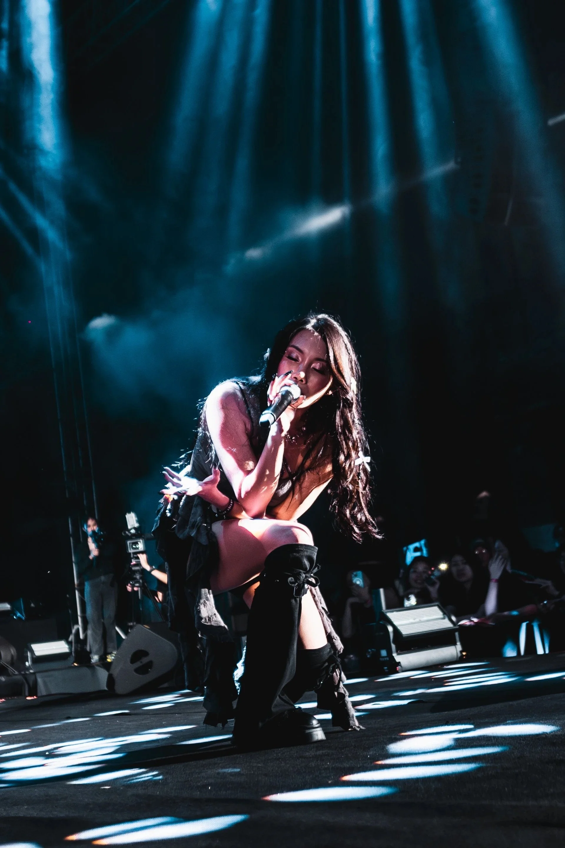 A female singer performs on stage under dramatic blue lighting, kneeling with a microphone in hand, with an audience and stage equipment visible in the background.