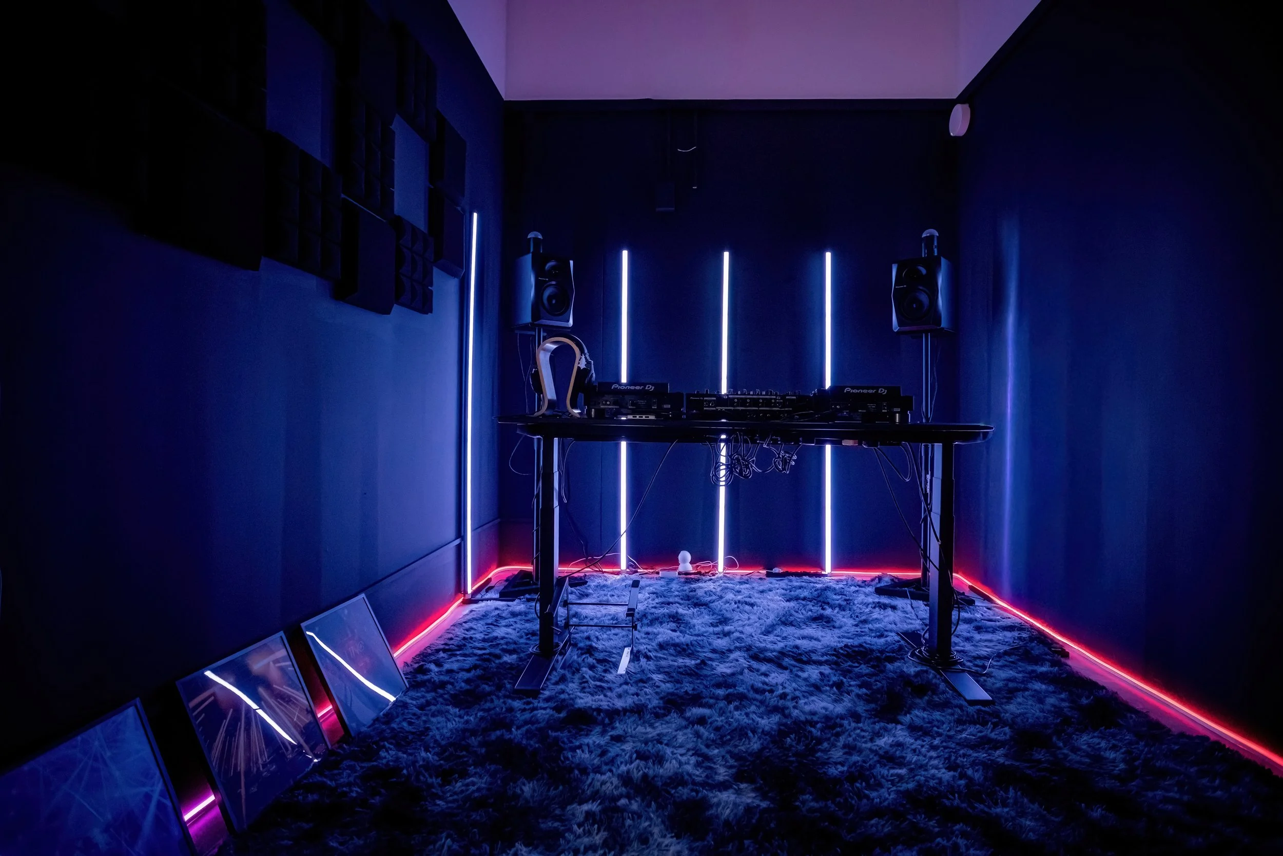 A dark music studio with blue and red LED lighting, a DJ table with Pioneer equipment, speakers, headphones, and framed artwork leaning against the wall.