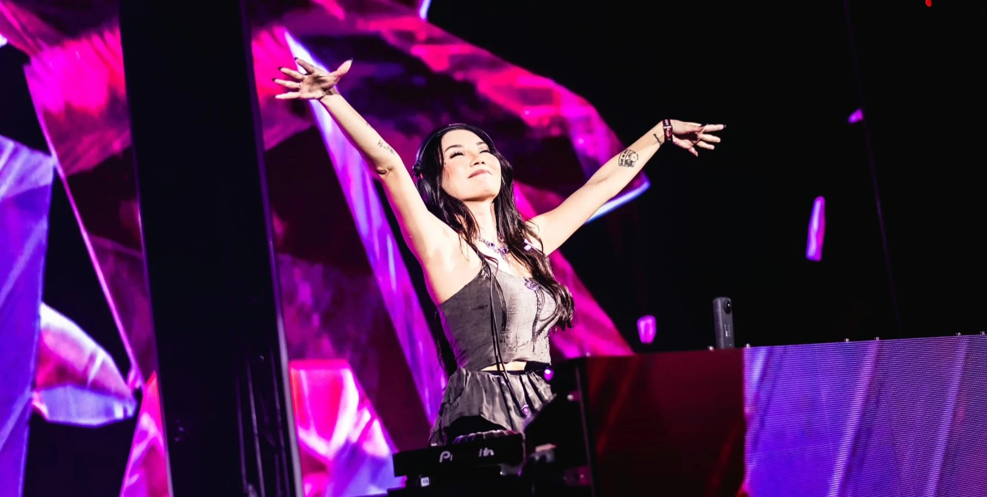 A female DJ with long dark hair and tattoos on her arms, wearing headphones, stands behind a DJ booth with her arms raised, smiling, during a live music performance with vibrant pink and purple stage lighting.